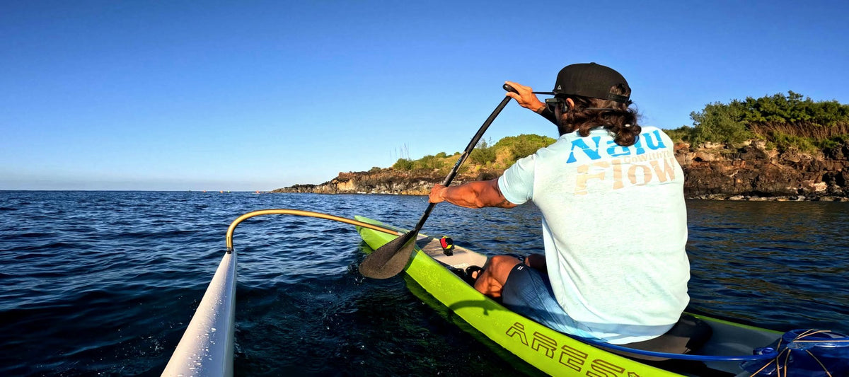 A paddler in an OC1 heads along the coast, seen from the ama of a second canoe