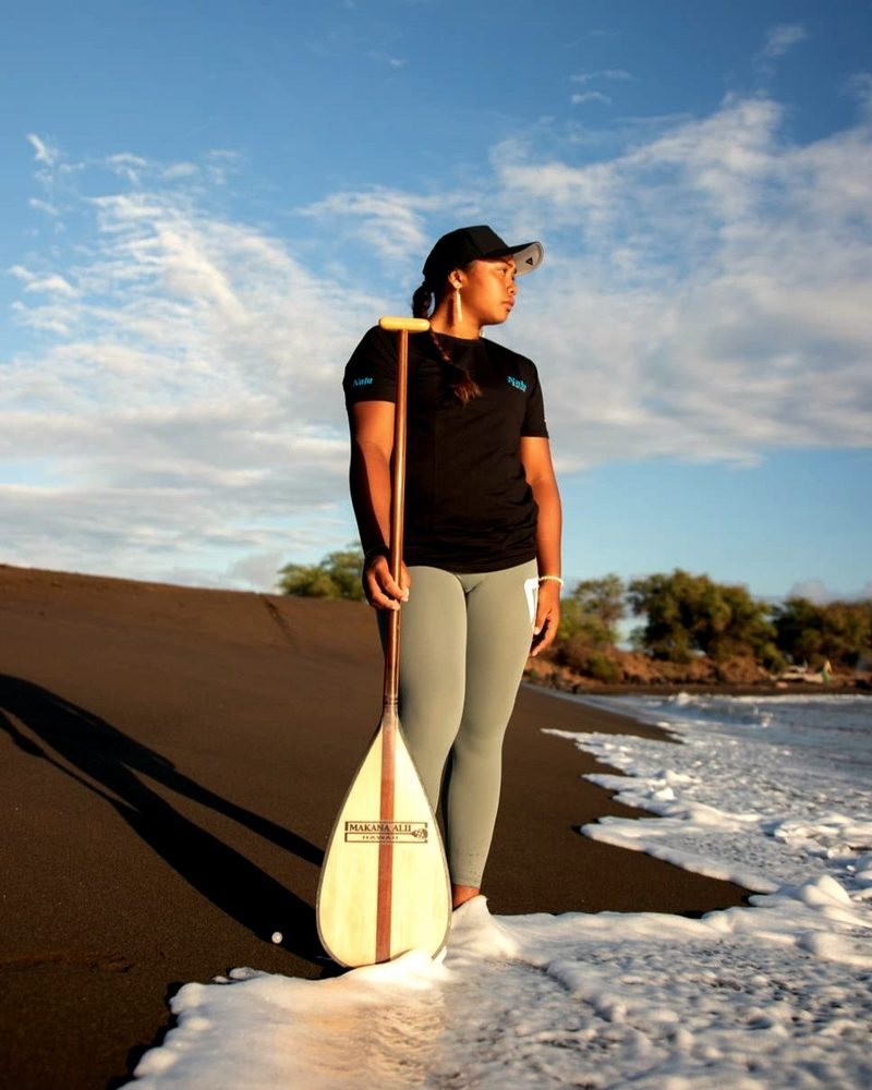 A paddler holds her paddle on a dark-sand beach at the water's edge